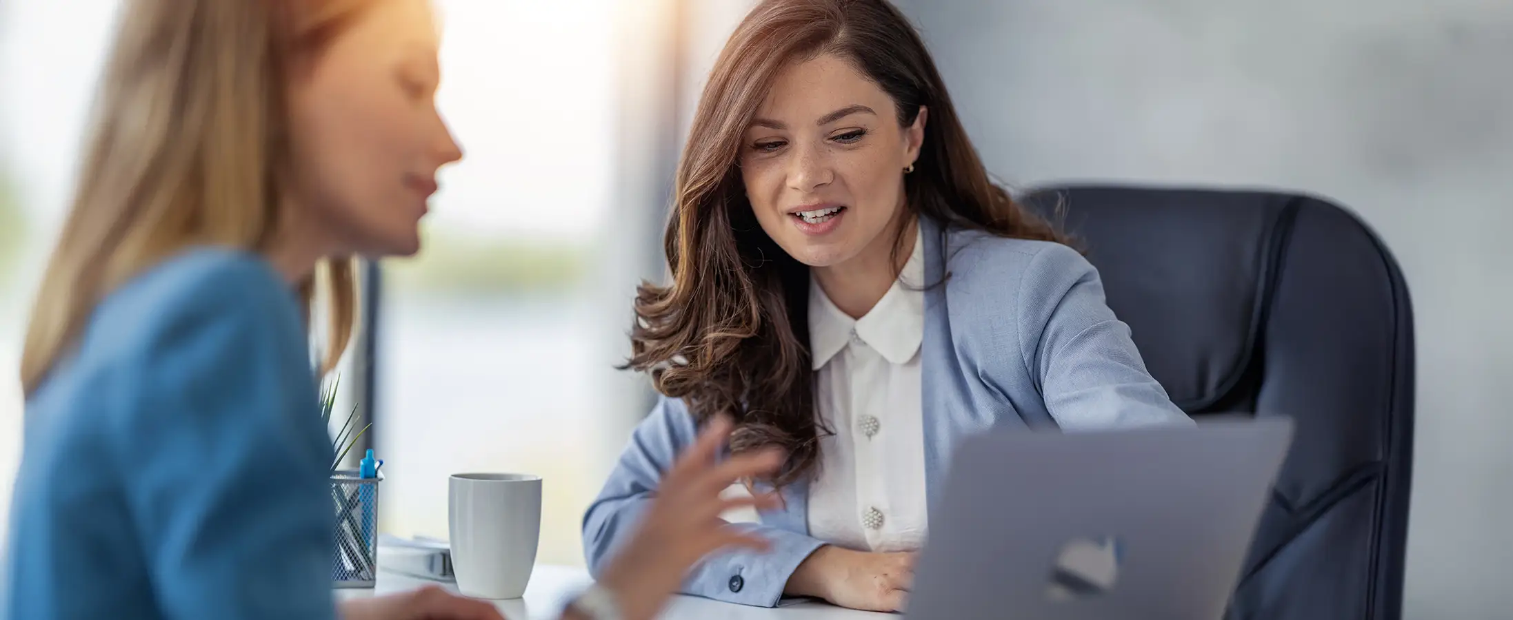 A woman in a blue blazer and white shirt points at a laptop screen while working with another woman in an office