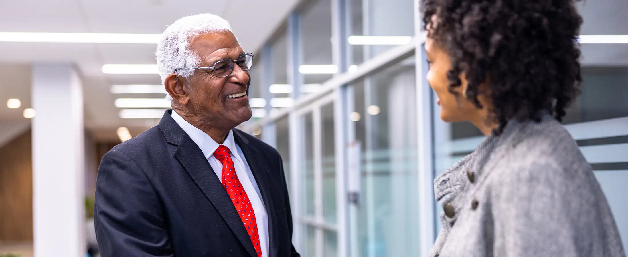 An older man wearing glasses and a suit and tie smiling and greeting a woman in a grey jacket in an office setting