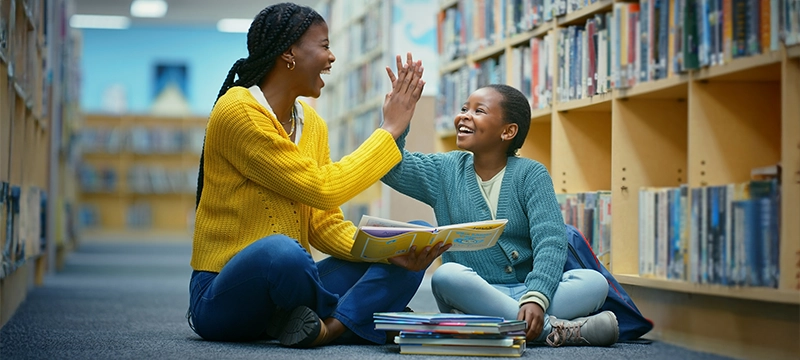 A young student high fives her smiling teacher as they sit in a library together with an open book, symbolizing that the FAR helped the girl with her reading abilities