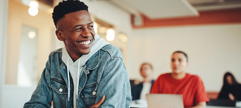 A male student in a denim jacket smiles in a brightly lit classroom, representing student success and self confidence