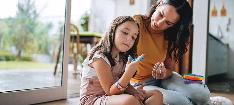 A woman site with her child in the living area of a home as the child (wearing a puzzle-patterned bracelet), who appears to have autism, plays with sensory tools to convey the impact of autism and the need for autism assessment.