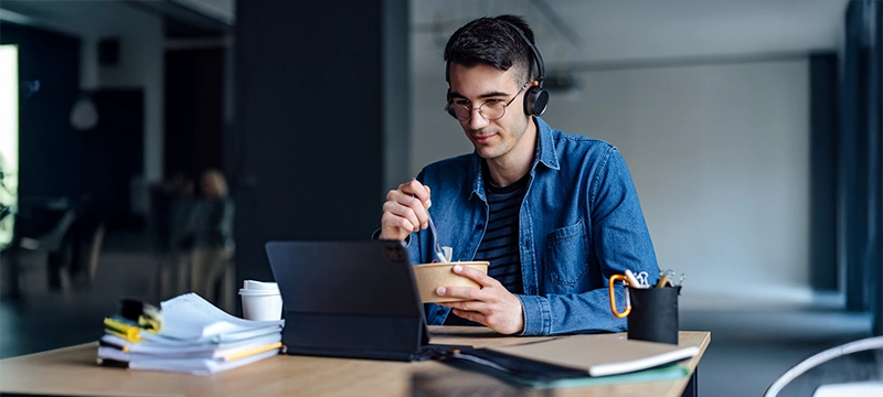 A man sits at a desk while eating and watching a webinar on a tablet with files stacked beside him, representing how busy psychologists can continue earning credits with a busy schedule with on-demand CE