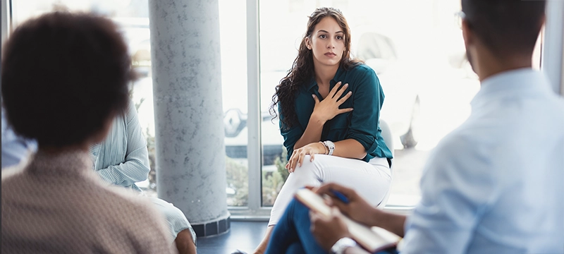 A group of adults sit in a circle facing an addiction professional with a clipboard and notepad, symbolizing addiction treatment and how personality affects addiction