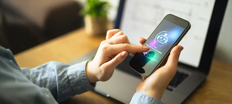 A person sitting at a desk holds up a phone with an image of an AI chatbot, representing using AI to save time